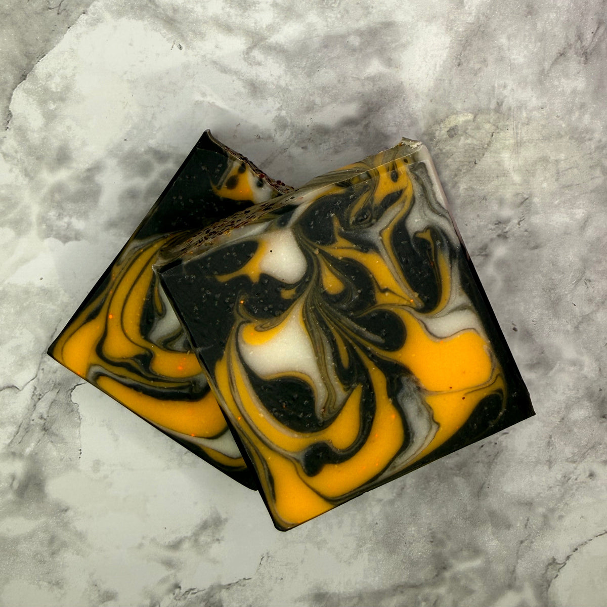 Two rectangular artisanal soaps with a marbled pattern of black, yellow, and white, arranged on a gray and white marble surface.