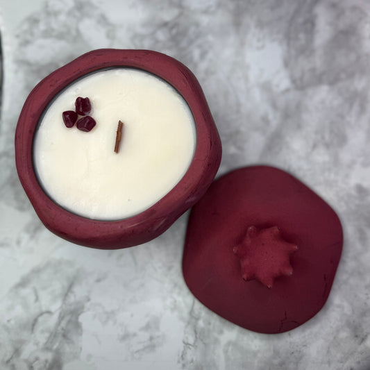 A small wax candle in a matte burgundy dish with its matching lid placed beside it on a light gray marble-patterned surface. The candle wax is off-white and the top is decorated with three small dark red buds and a single thin wick.
