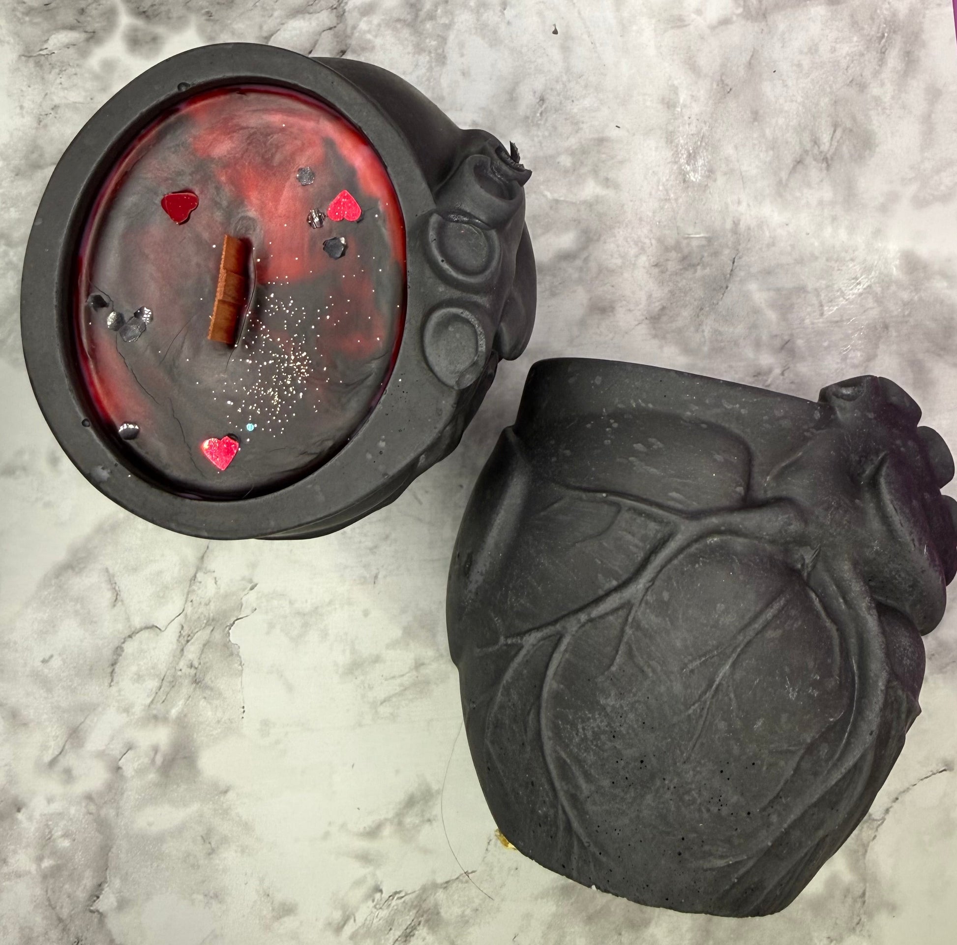 A black anatomical heart-shaped candle shown open, with red wax inside and small red heart details, alongside the matching black heart-shaped lid, on a gray marble-patterned surface.
